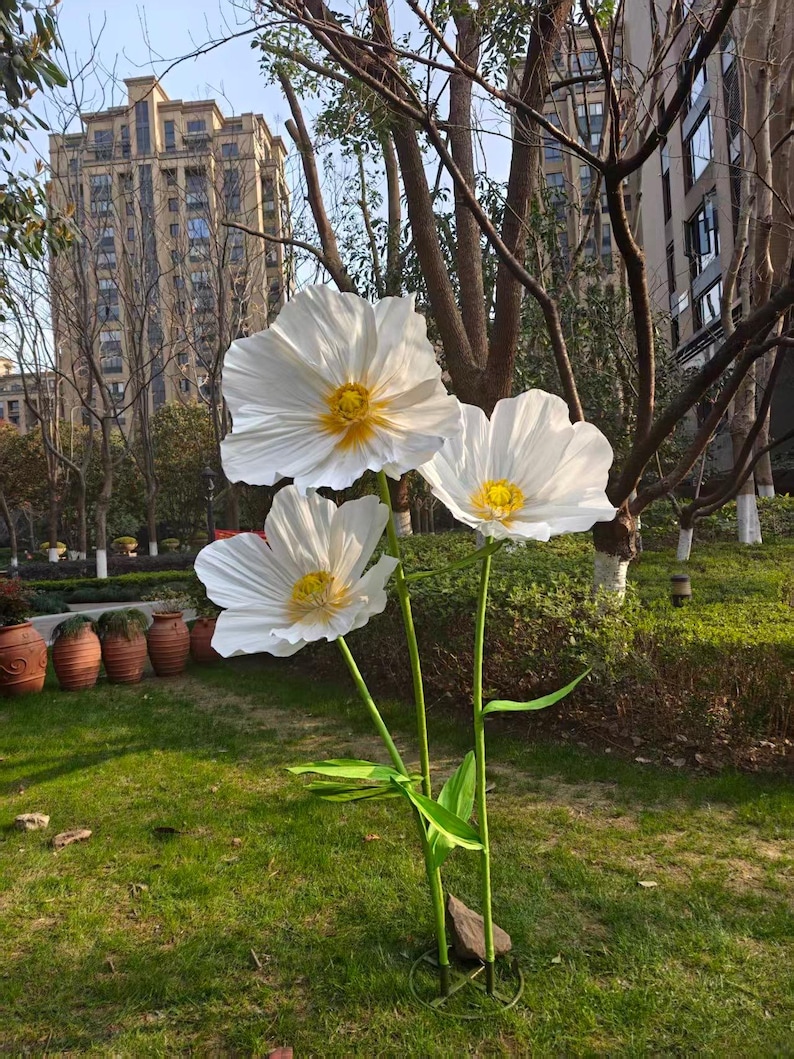Peut inclure: Trois grandes sculptures de fleurs blanches en m&eacute;tal avec des centres jaunes se dressent dans une zone herbeuse. Les fleurs ont des p&eacute;tales d&eacute;licats et froiss&eacute;s et sont soutenues par des tiges et des feuilles vertes. L'arri&egrave;re-plan comprend des arbres et un grand b&acirc;timent.