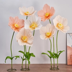 Peut inclure: Un ensemble de grandes fleurs en papier aux tons p&ecirc;che et blanc, avec des centres jaunes. Les fleurs ont des tiges et des feuilles vertes, et sont dispos&eacute;es de mani&egrave;re d&eacute;corative. Les fleurs sont sur un socle en m&eacute;tal.