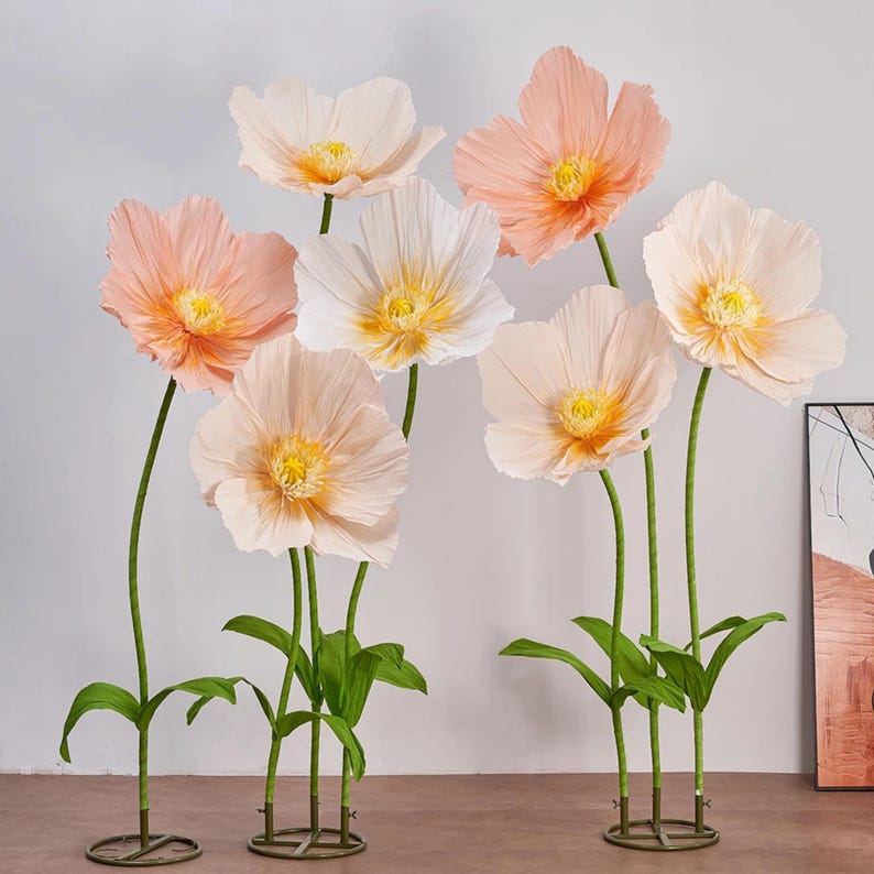 Peut inclure: Un ensemble de grandes fleurs en papier aux tons p&ecirc;che et blanc, avec des centres jaunes. Les fleurs ont des tiges et des feuilles vertes, et sont dispos&eacute;es de mani&egrave;re d&eacute;corative. Les fleurs sont sur un socle en m&eacute;tal.