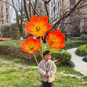 Peut inclure: Un enfant se tient devant une grande installation artistique ext&eacute;rieure de trois fleurs orange et jaune. Les fleurs ont un centre sombre et sont mont&eacute;es sur une tige verte. L'enfant porte une veste beige, un pantalon noir et des baskets bleues et blanches.