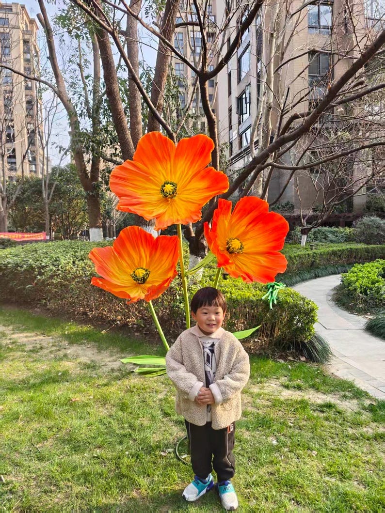 Peut inclure: Un enfant se tient devant une grande installation artistique ext&eacute;rieure de trois fleurs orange et jaune. Les fleurs ont un centre sombre et sont mont&eacute;es sur une tige verte. L'enfant porte une veste beige, un pantalon noir et des baskets bleues et blanches.
