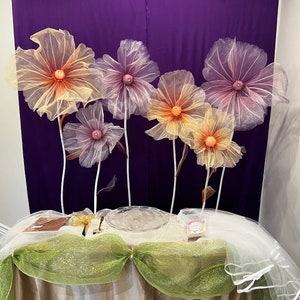 Puede incluir: Una mesa decorada con un mantel blanco, tul verde y blanco y un plato de vidrio transparente. Seis grandes flores delicadas de papel de seda en tonos rosa y amarillo est&aacute;n dispuestas en el fondo.
