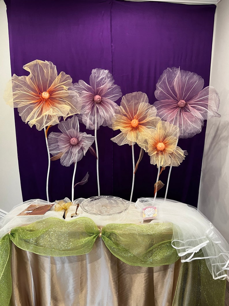Puede incluir: Una mesa decorada con un mantel blanco, tul verde y blanco y un plato de vidrio transparente. Seis grandes flores delicadas de papel de seda en tonos rosa y amarillo est&aacute;n dispuestas en el fondo.