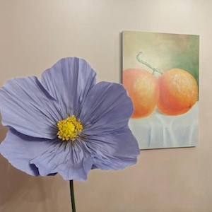 Peut inclure: Une grande fleur en papier violet clair avec un centre jaune et une tige verte. Une peinture de deux oranges sur un tissu blanc est accroch&eacute;e au mur en arri&egrave;re-plan.