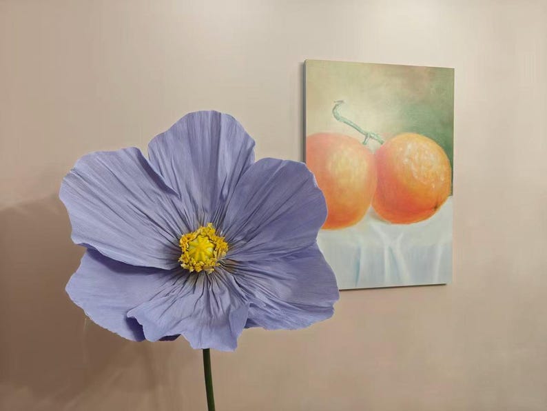 Peut inclure: Une grande fleur en papier violet clair avec un centre jaune et une tige verte. Une peinture de deux oranges sur un tissu blanc est accroch&eacute;e au mur en arri&egrave;re-plan.