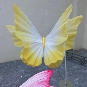 Puede incluir: Una gran decoración de mariposa amarilla y blanca con un centro amarillo. La mariposa está unida a un alambre delgado y está de pie sobre un soporte plateado.