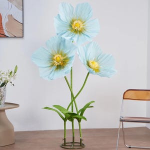 Peut inclure: Trois grandes fleurs de papier bleu clair avec des centres jaunes et des tiges vertes. Les fleurs artificielles ont des feuilles vertes et sont dispos&eacute;es dans une base m&eacute;tallique d&eacute;corative, cr&eacute;ant une exposition florale saisissante.