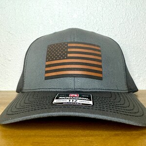 May include: A gray and black trucker hat with a brown leather American flag patch on the front. The hat has an adjustable strap and the text "Richardson 112 Original Trucker Adjustable" on the back.