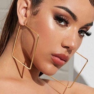 May include: A close-up shot of a person wearing large, gold square hoop earrings. The earrings are a simple, geometric design, and the gold color is bright and shiny. The person has dark hair and is wearing makeup.