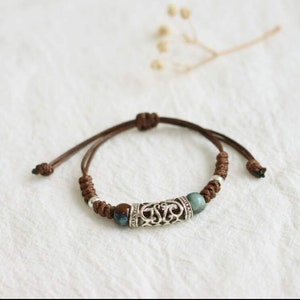 May include: A brown cord bracelet with a silver charm and blue and green beads.
