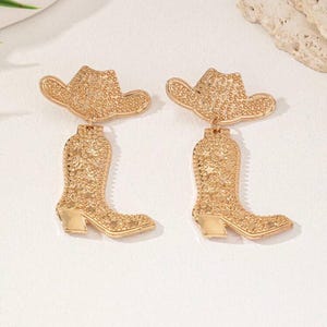 May include: A pair of gold-tone earrings featuring a cowboy boot and hat design. The earrings are made of metal and have a textured surface.
