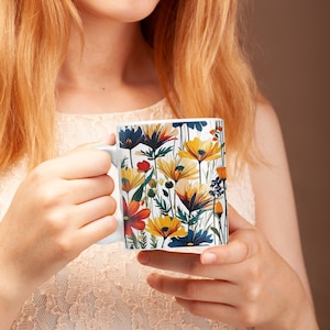 May include: White ceramic mug featuring a colorful floral design with orange, yellow, and blue flowers. The mug has a white handle and is held by a person. The floral pattern includes various flower types and green foliage.
