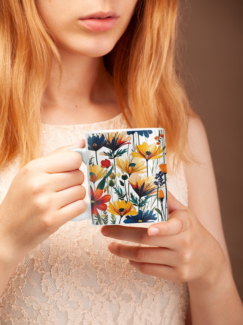 May include: White ceramic mug featuring a colorful floral design with orange, yellow, and blue flowers. The mug has a white handle and is held by a person. The floral pattern includes various flower types and green foliage.
