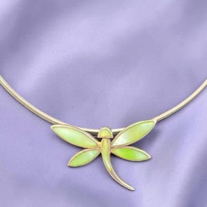 May include: A gold-toned necklace featuring a dragonfly pendant. The dragonfly has light green enamel wings and a matching body. The necklace has a simple, curved design, resting on a lavender surface.