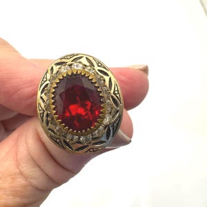 May include: An oval-shaped ring featuring a large, faceted, red gemstone. The ring has a gold-toned band with a decorative black pattern and a row of small, clear stones around the red stone. The ring is held by a hand.