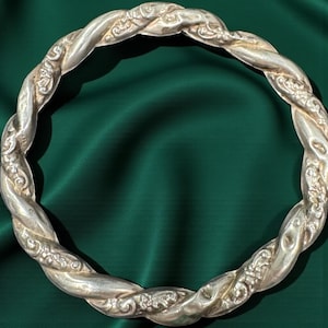 May include: A silver-colored, twisted metal bangle bracelet. The bracelet has a braided design with ornate floral detailing. The background is a dark green fabric, highlighting the bracelet's intricate design and metallic sheen.