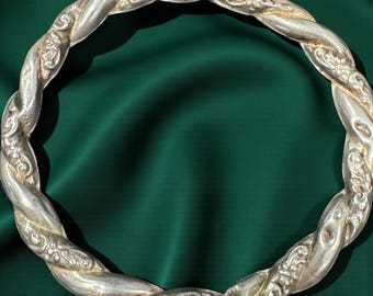 May include: A silver-colored, twisted metal bangle bracelet. The bracelet has a braided design with ornate floral detailing. The background is a dark green fabric, highlighting the bracelet's intricate design and metallic sheen.