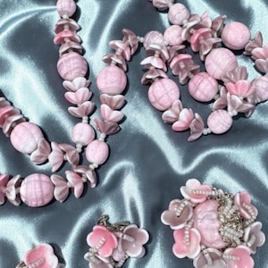 May include: A vintage jewelry set featuring necklaces, earrings, and a brooch. The set is primarily pink, with flower and bead details. The necklaces have large, round beads and flower-shaped accents. The earrings and brooch also feature flower designs.