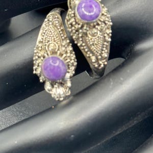 May include: Two ornate silver rings with a purple gemstone center. The rings have detailed filigree and beadwork designs around the gemstones. The rings are set against a dark background.