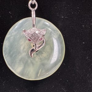 May include: A silver pendant necklace with a jade green circle charm featuring a silver fox with clear and red gemstone accents.