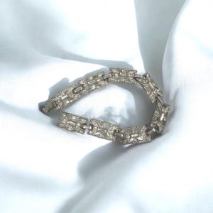 May include: Silver-toned bracelet with rectangular links, each adorned with small, sparkling rhinestones. The bracelet lies on a white, silky fabric, catching the light and highlighting its intricate design. A classic piece of jewelry.