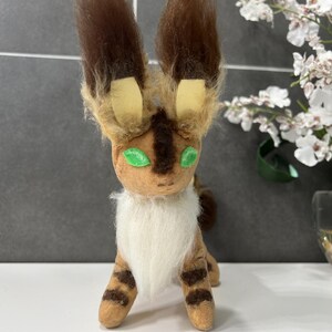 May include: A handmade plush creature with brown and tan fur, green eyes, and large, upright ears. The plush has a white chest and striped legs, resembling a stylized animal or mythical being. The toy is standing on a white surface.