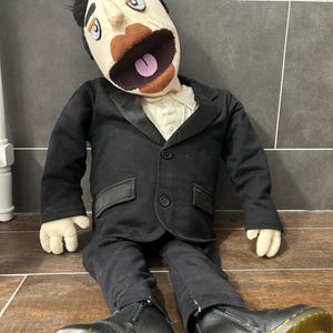 May include: A handmade puppet doll with a fabric face, black hair, and a black suit. The doll is wearing a white collared shirt and black pants. It is wearing black leather boots with yellow soles.