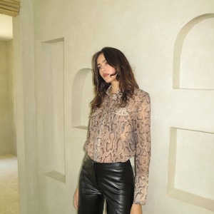 Könnte beinhalten: Eine Frau trägt eine langärmelige Bluse mit Schlangenhautmuster, einen hohen Kragen und eine schwarze Lederhose.