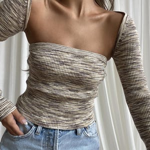 May include: A beige and brown striped long-sleeve top with a unique open-shoulder design. The top is paired with light blue denim jeans. The fabric has a ribbed texture.