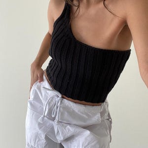 May include: A black one-shoulder knitted crop top with a ribbed texture. The top is paired with white cargo pants.