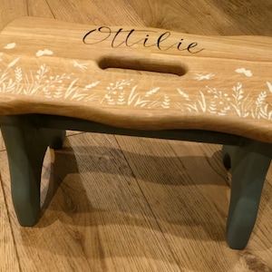 May include: A wooden step stool with a light blue base and a natural wood top. The top is decorated with a white floral design and the name "Ottilie" is engraved in black lettering.