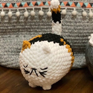May include: A crocheted stuffed cat toy with a white body, black and orange patches, and a white tail. The cat has a black embroidered face with closed eyes.