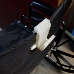 May include: A white, 3D-printed clip is attached to the black fabric of a wheelchair. The clip has a rectangular body and two curved hooks. The wheelchair has a black seat and backrest, with a metal frame and wheels.