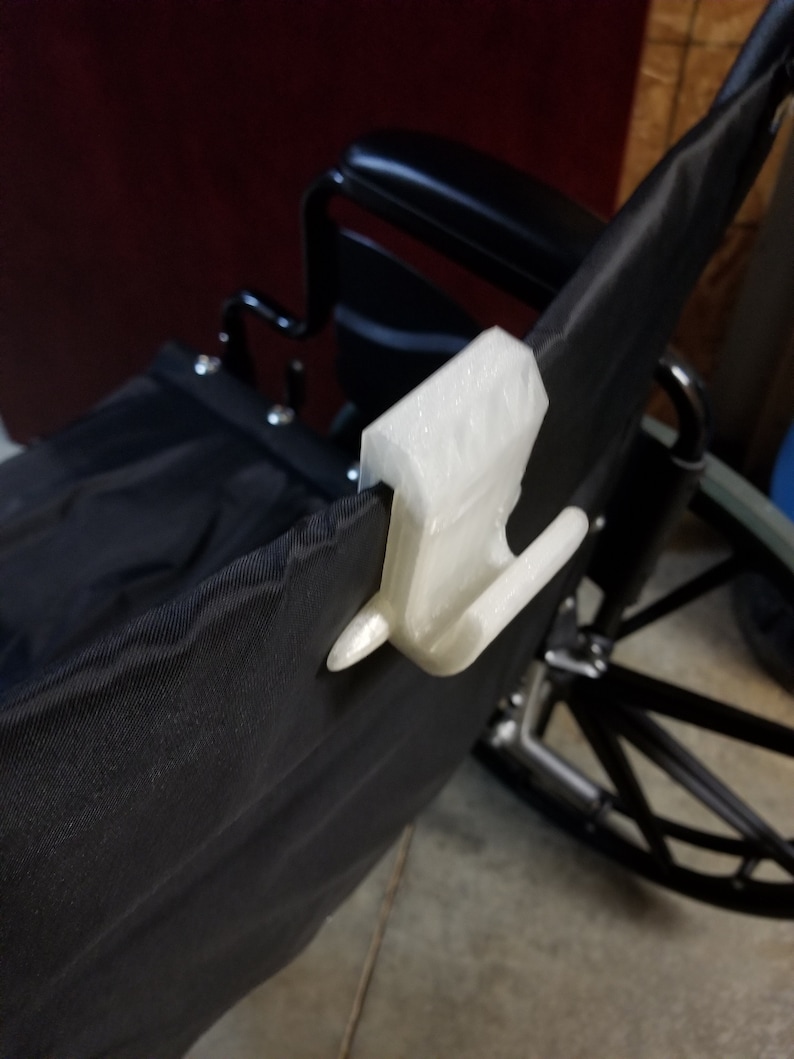 May include: A white, 3D-printed clip is attached to the black fabric of a wheelchair. The clip has a rectangular body and two curved hooks. The wheelchair has a black seat and backrest, with a metal frame and wheels.