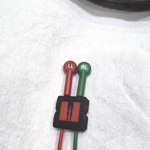May include: Red and green medical device with the text "LL" and "RL" on the ends. The device is connected to a black square with a red center and a black graphic. The device is on a white surface.