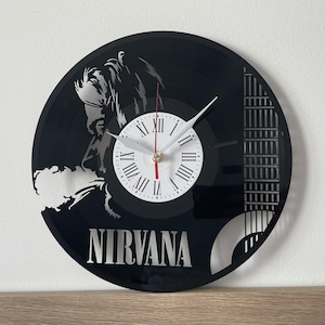 Può includere: Orologio in vinile nero con la silhouette di una persona e la parola "NIRVANA" stampata sotto. Il quadrante dell'orologio è bianco con numeri romani neri.