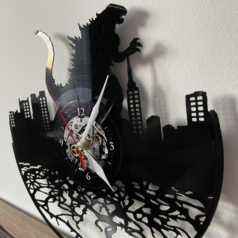 May include: Black vinyl record clock featuring a silhouette of Godzilla stomping on a city skyline. The clock face has Roman numeral markings and a gear-like design.