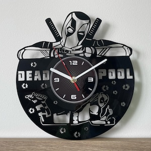 Pode incluir: Relógio de vinil preto com uma silhueta do Deadpool e as palavras "DEADPOOL" no mostrador. Os ponteiros do relógio são brancos e vermelhos.