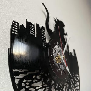 May include: Black vinyl record clock featuring a silhouette of Godzilla destroying a city. The clock face is visible with black hands and a gold gear mechanism.