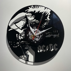 Könnte beinhalten: Schwarze Schallplattenuhr mit einer weißen Silhouette einer Person, die Gitarre spielt, und dem Text "AC/DC" darunter. Das Zifferblatt der Uhr ist ein kleinerer Kreis mit römischen Ziffern.