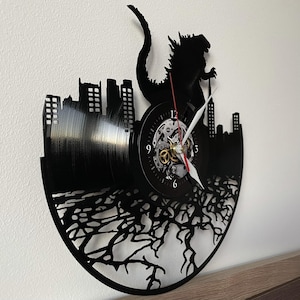 May include: Black vinyl record clock featuring a silhouette of Godzilla destroying a city. The clock face is white with black numbers and hands.
