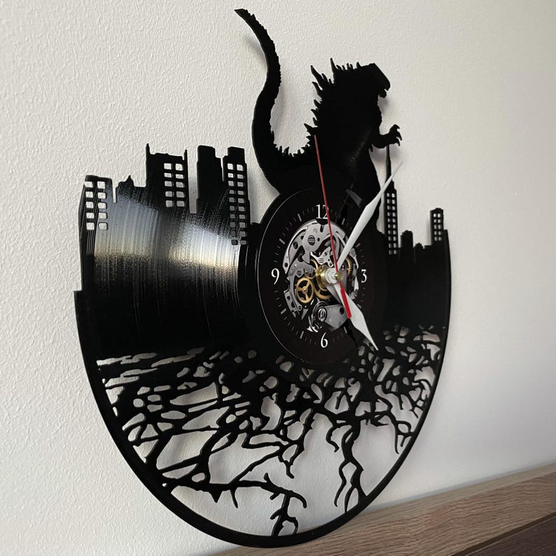 May include: Black vinyl record clock featuring a silhouette of Godzilla destroying a city. The clock face is white with black numbers and hands.