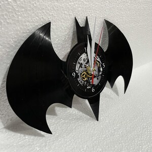 K&ouml;nnte beinhalten: Eine schwarze Schallplatte in Form einer Fledermaus mit einem Zifferblatt in der Mitte. Das Zifferblatt hat ein skelettartiges Design mit sichtbaren Zahnr&auml;dern und einem roten Sekundenzeiger.