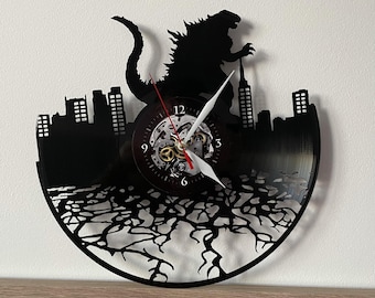 May include: Black vinyl record clock featuring a silhouette of Godzilla stomping on a city skyline. The clock face is visible with Roman numeral markings and a gear-like design.