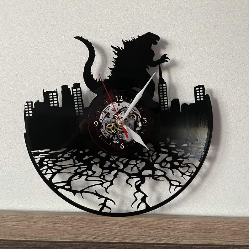 May include: Black vinyl record clock featuring a silhouette of Godzilla stomping on a city skyline. The clock face is visible with Roman numeral markings and a gear-like design.