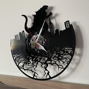 May include: Black vinyl record clock featuring a silhouette of Godzilla destroying a city skyline. The clock face is visible with Roman numeral markings and a white hand.