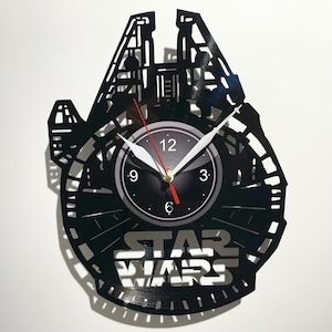 May include: A black vinyl record clock featuring a silhouette of the Millennium Falcon from Star Wars. The clock face is black with white numbers and hands. The words "Star Wars" are printed in white below the clock face.