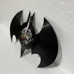K&ouml;nnte beinhalten: Eine schwarze Schallplatte in Form des Batman-Logos mit einem Zifferblatt in der Mitte. Das Zifferblatt hat einen schwarzen Hintergrund mit wei&szlig;en Zahlen und einem silbernen und goldenen Uhrwerk.