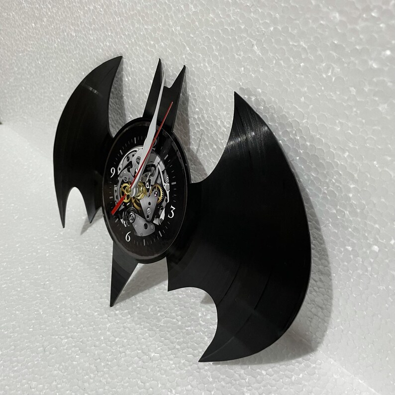 K&ouml;nnte beinhalten: Eine schwarze Schallplatte in Form des Batman-Logos mit einem Zifferblatt in der Mitte. Das Zifferblatt hat einen schwarzen Hintergrund mit wei&szlig;en Zahlen und einem silbernen und goldenen Uhrwerk.