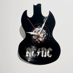 Könnte beinhalten: Schwarze, gitarrenförmige Wanduhr aus einer Schallplatte. Die Uhr hat ein sichtbares Zahnradwerk, weiße Zeiger und den Bandnamen "AC/DC" in Weiß. Der Sekundenzeiger ist rot.